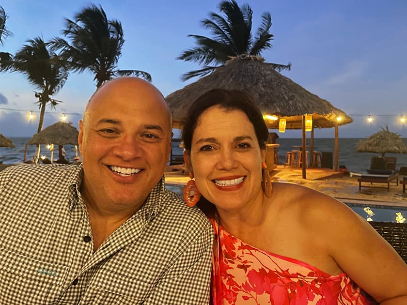 A smiling couple poses together outdoors at dusk, with palm trees and beach huts in the background. A smiling couple poses together outdoors at dusk, with palm trees and beach huts in the background.