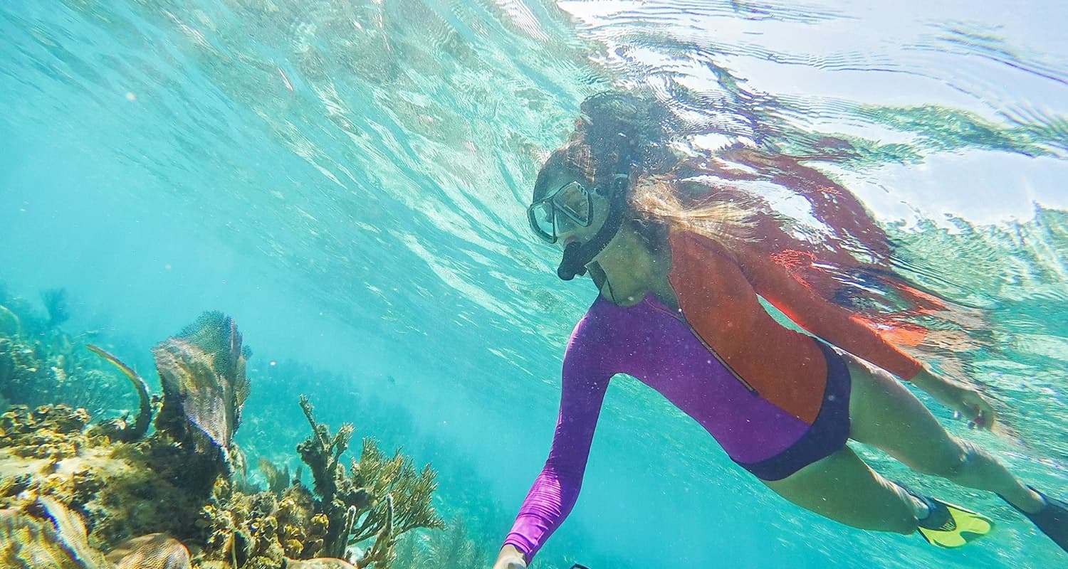 A woman in a red and purple scuba top and yellow fins snorkels in the clear blue water near underwater plant life.