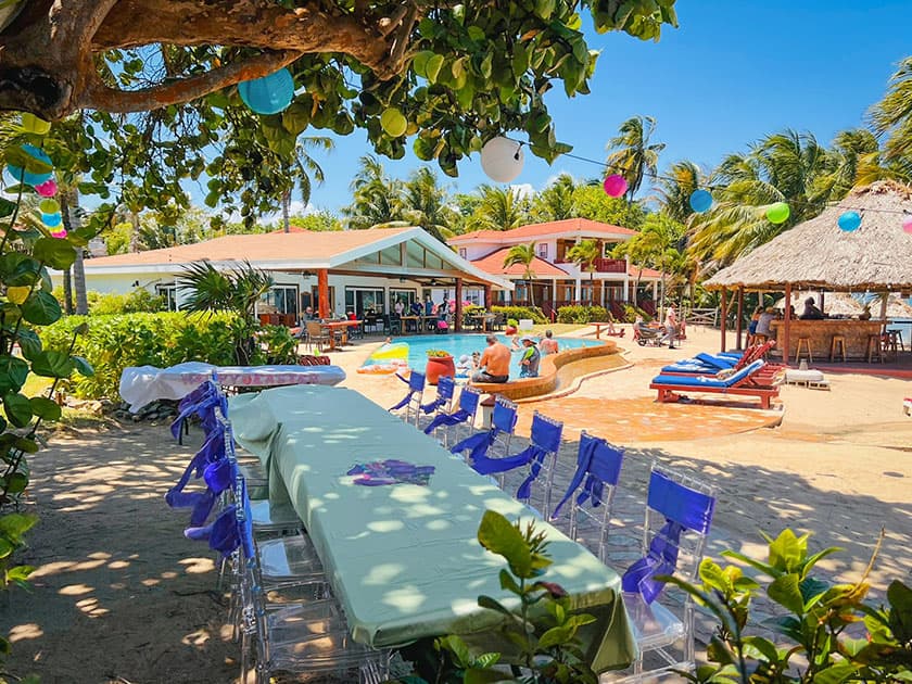 A tropical resort scene featuring a long table, lounge chairs, a pool, and colorful decorations amid palm trees. A tropical resort scene featuring a long table, lounge chairs, a pool, and colorful decorations amid palm trees.