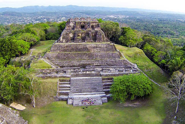 Aerial view of an ancient stone pyramid surrounded by lush greenery and distant hills.
