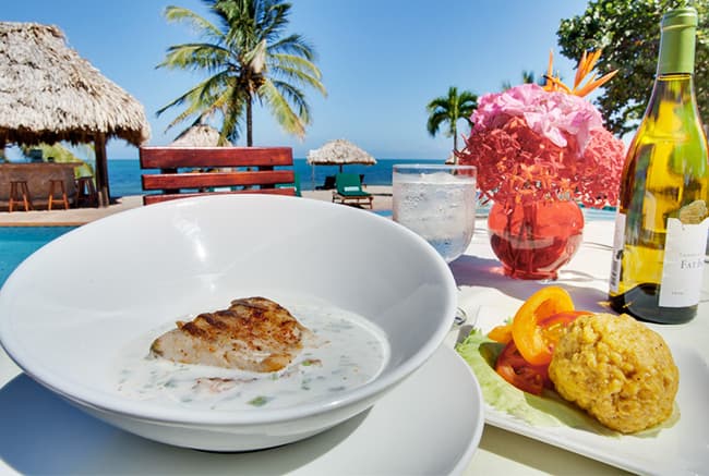 A gourmet meal featuring grilled fish and sides, with a tropical beach and palm trees in the background.