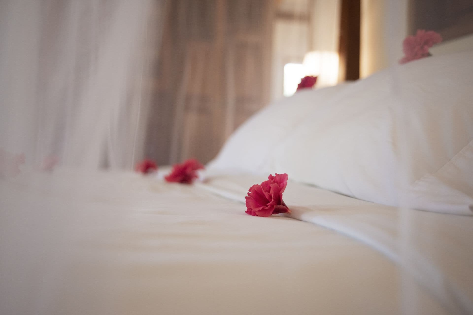 A spacious bedroom with a four-poster bed covered in white sheets. The bed has sheer white curtains hanging from the canopy. A ceiling fan hangs from the center of the room, and there is an air conditioning unit in the background. Nightstands with lamps flank the bed, and a wooden bench sits at the foot. A small sofa sits in the corner of the room, and a coffee table is placed between the sofa and the bed. The room has a tiled floor and a wooden closet door. A colorful painting hangs on the wall behind the bed.