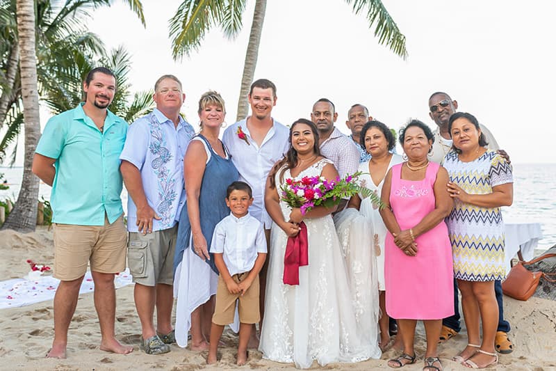 A bride in a white dress holds a bouquet while posing with her family and friends on a beach.