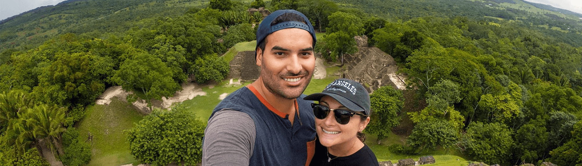 Man and woman in ball caps and sunglasses standing above a mayan ruin taking a selfie.