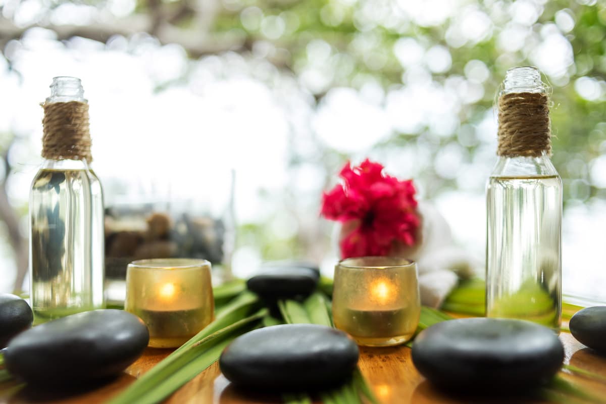 The image shows a serene spa setting with two bottles of essential oil, a red flower, two candles, and several smooth stones on a wooden table. The background is blurred and suggests a lush, green setting.