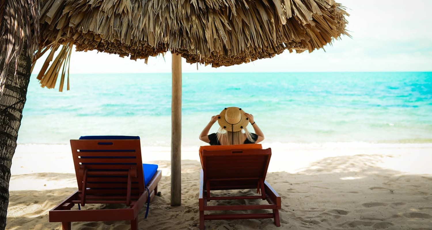 A single woman in a straw hat sitting in one of two beach chairs under thatch umbrella alone, on beach look at the ocean.