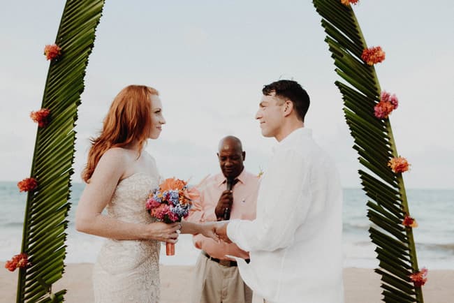 A couple exchanges vows under a floral arch on the beach, with an officiant in the background.