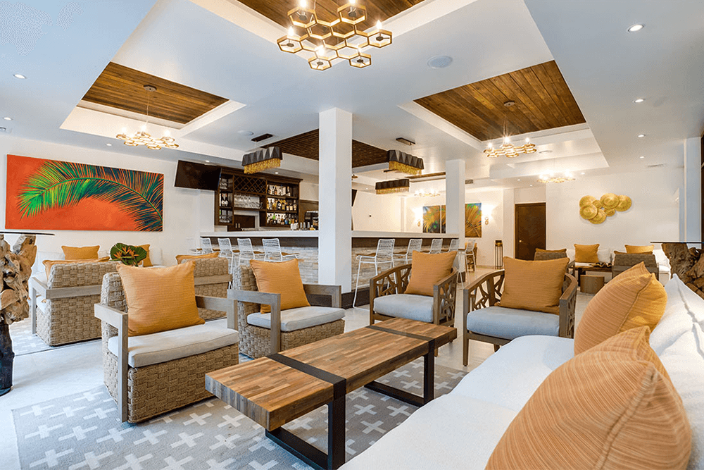 A spacious and modern hotel lobby with a high ceiling and a large, abstract painting on the wall. Comfortable seating arrangements, including sofas, armchairs, and bar stools, are arranged around a central coffee table. A bar area with a counter and stools is visible in the background.
