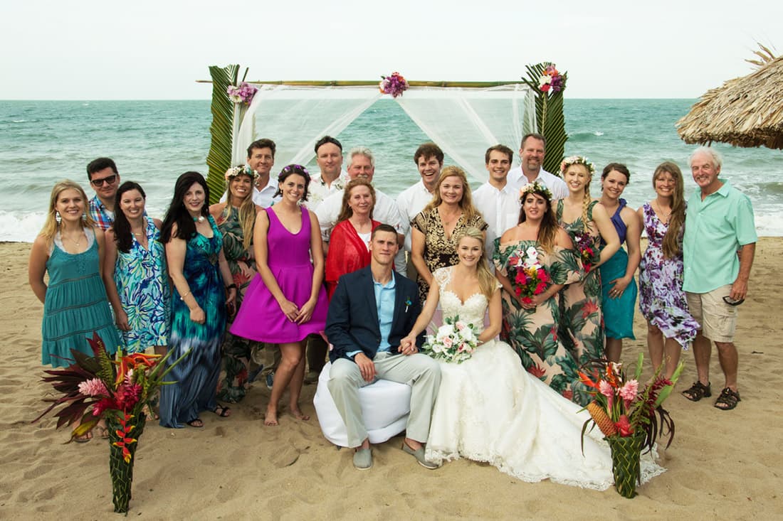 Handsome bride and groom pose on the beach, surrounded by smiling friends and family under a decorated wedding arch.