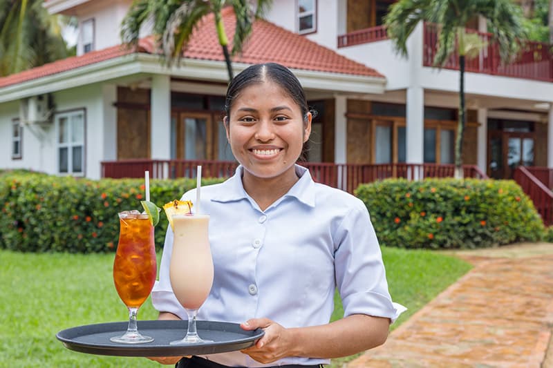 A smiling server presents two colorful cocktails on a tray outside a hospitality venue. A smiling server presents two colorful cocktails on a tray outside a hospitality venue.