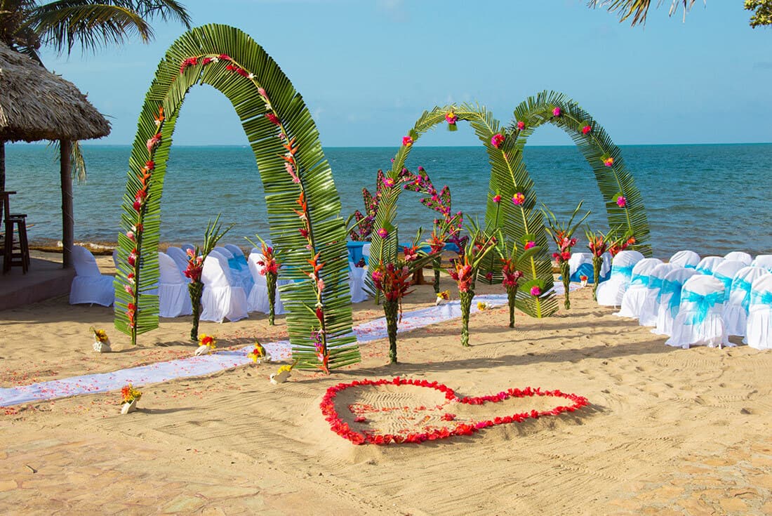 Tropical beach wedding ceremony setup with palm leaf arches, colorful flowers, and white chairs.