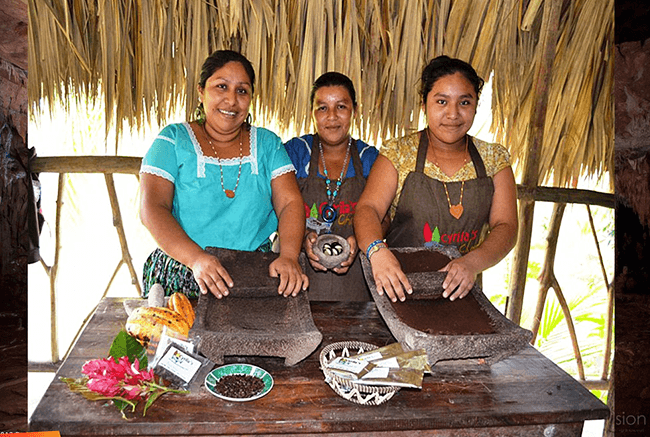 Three women stand behind a wooden table with traditional tools and ingredients, smiling in a tropical setting.