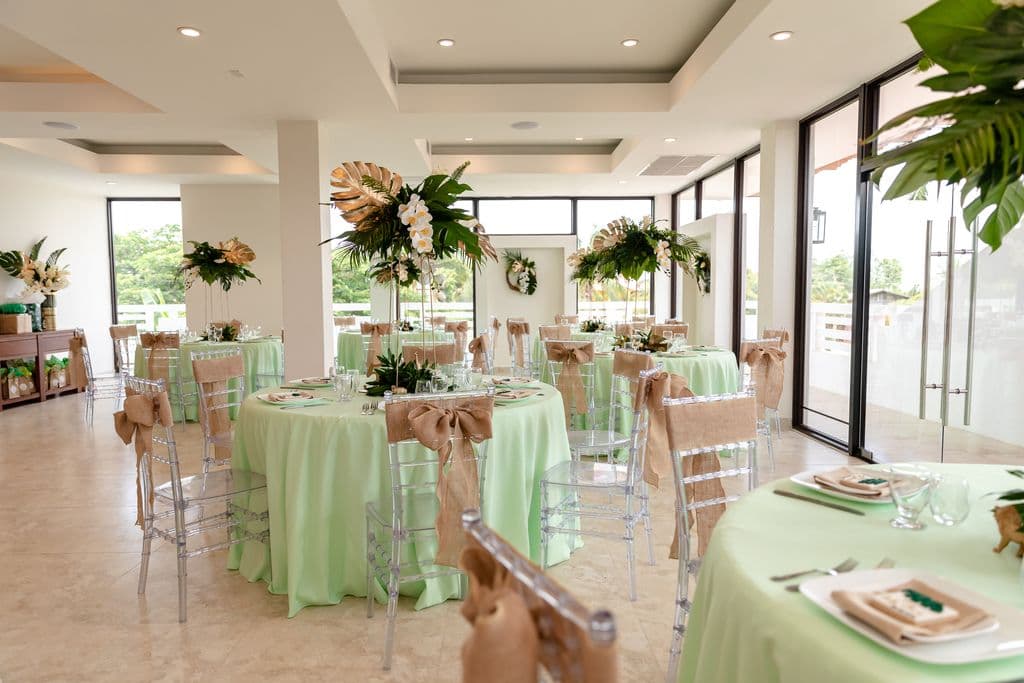 A beautifully decorated event space with a tropical theme. The room has white walls and large windows overlooking a green outdoor area. The tables are round and covered in light green tablecloths with burlap bows on the chairs. Tall floral centerpieces with tropical flowers and leaves adorn the tables. There are several potted plants around the room, adding to the tropical atmosphere.