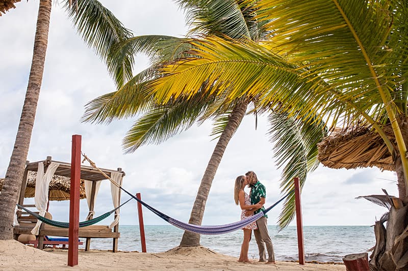 A couple shares a romantic kiss under palm trees on a beach with a hammock nearby. A couple shares a romantic kiss under palm trees on a beach with a hammock nearby.
