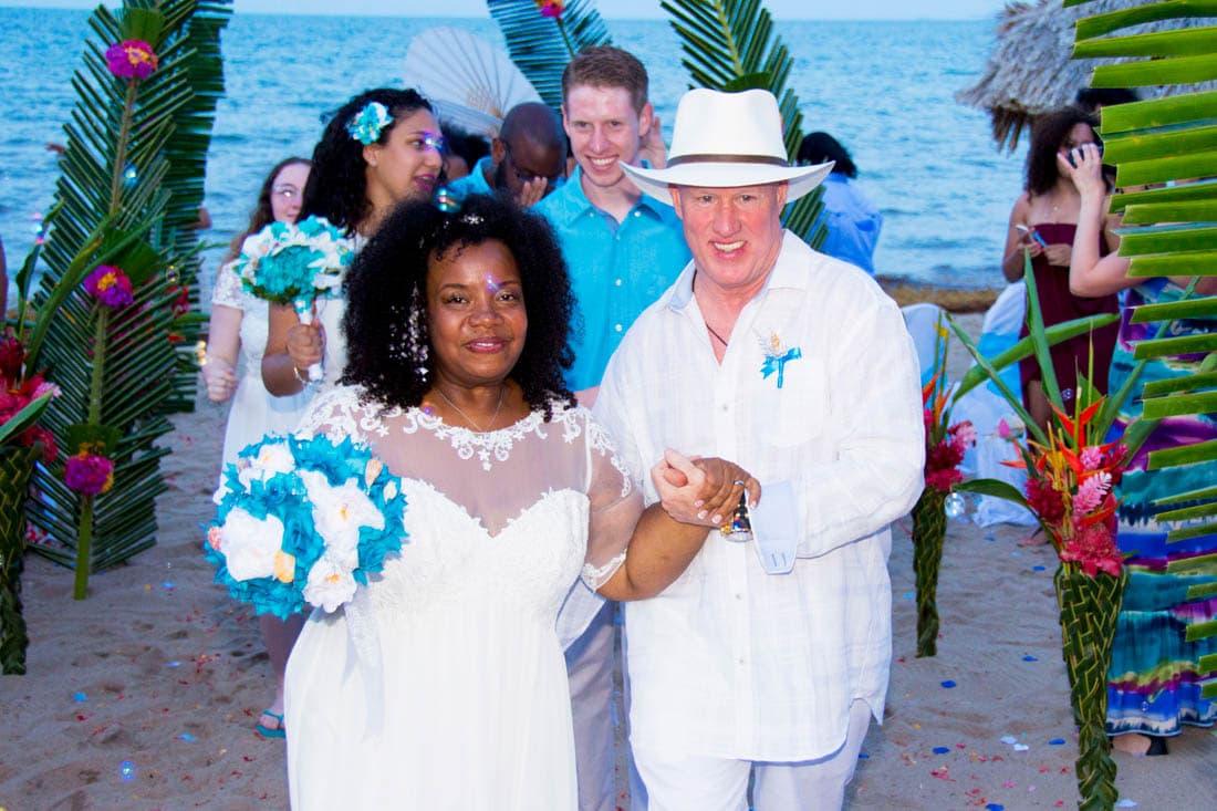 A happy couple walk through a wedding arch followed by the wedding party dressed in teal and white.