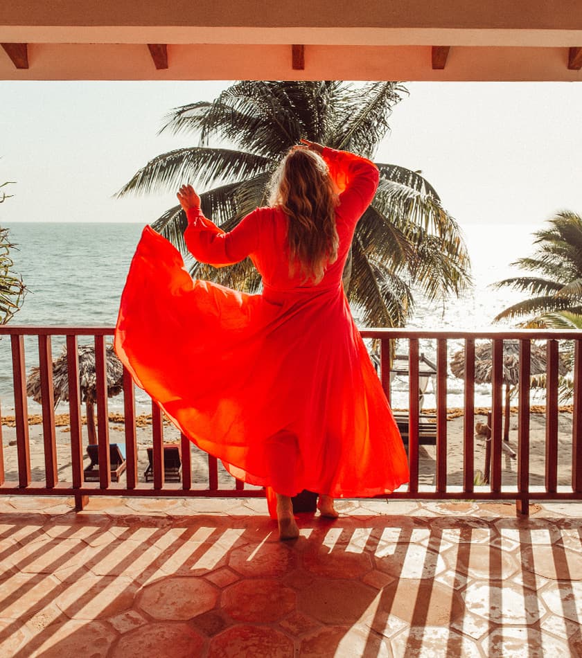 A person in a flowing red dress stands on a balcony overlooking the ocean and palm trees.