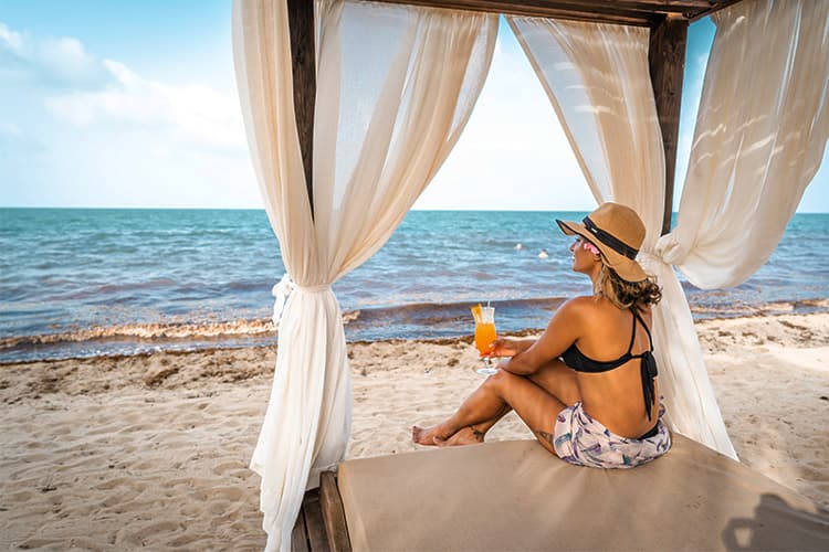 A woman in a swimsuit relaxes on a beach cabana, holding a cocktail and enjoying the view of the ocean.