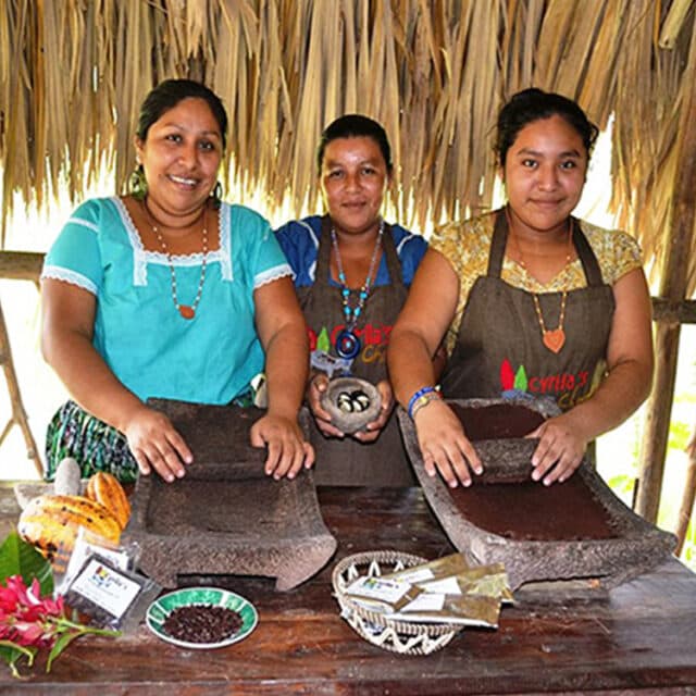Three ladies in colorful clothing stand before two metates, grinding chocolate beans.