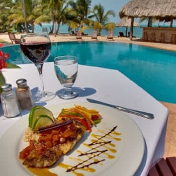 A plate of grilled fish with colorful vegetables and a side of rice is served on a table beside a swimming pool. A glass of red wine and a glass of water are also on the table. The background shows a thatched-roof bar and a beach.