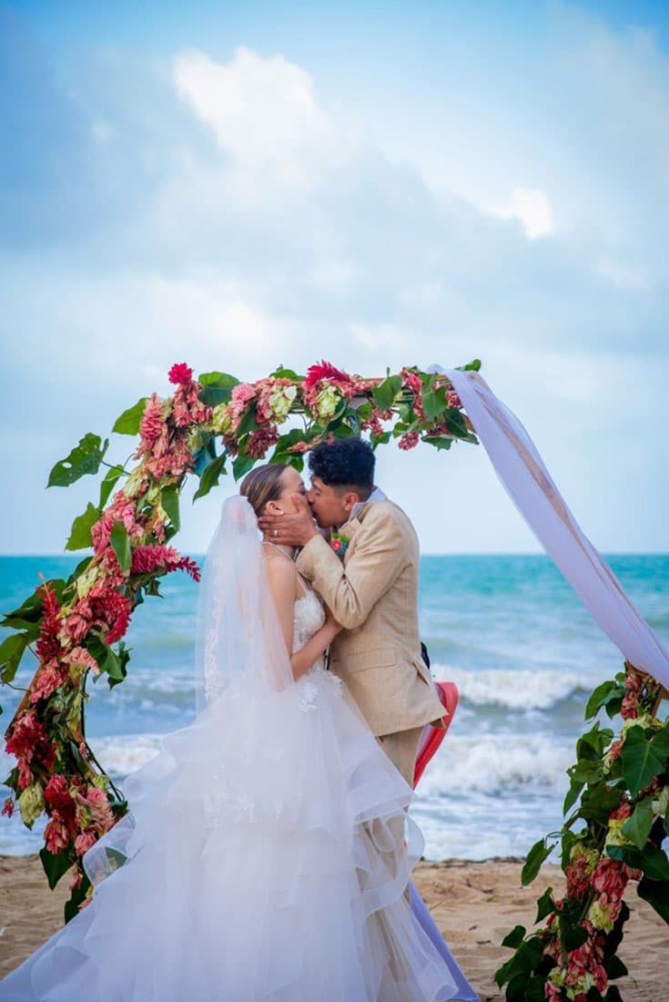 A young bride and groom kiss passionately on the beach under a wedding arch decked out with assorted pink and green florals.