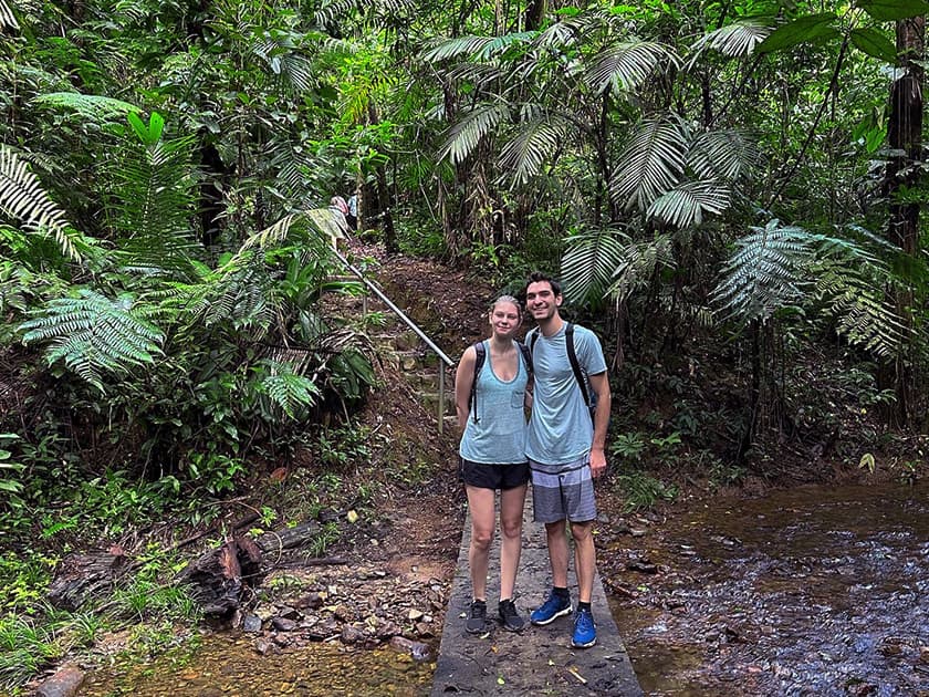 A couple stands on a stone path in a lush, green forested area near a stream.
