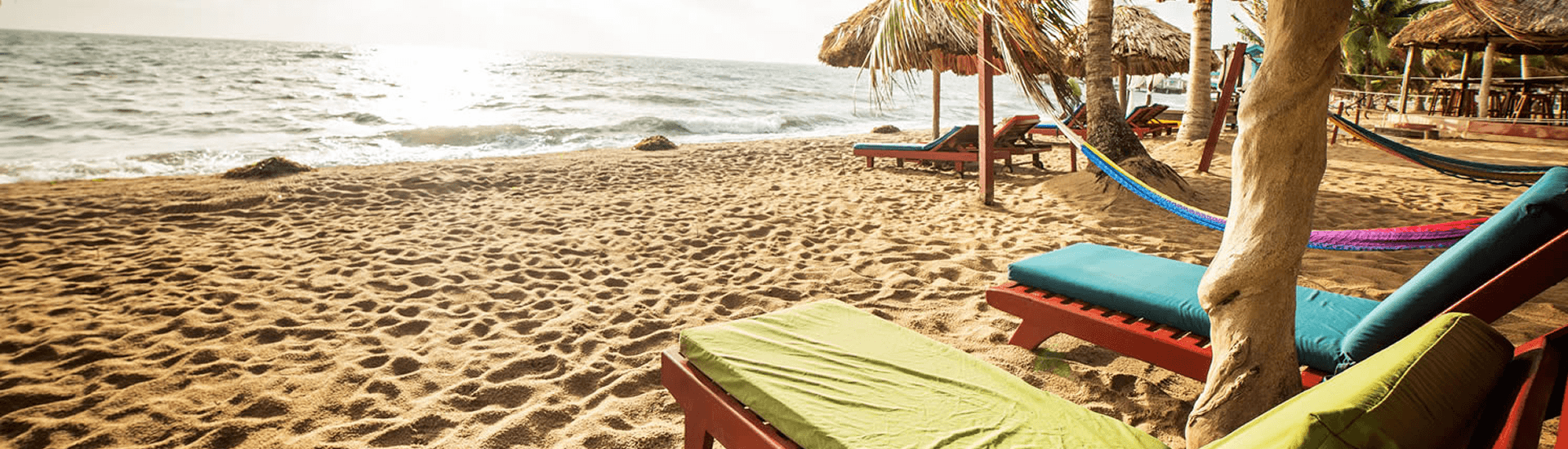 A serene beach scene with lounge chairs, hammocks, and thatched umbrellas. The sun shines brightly over the calm ocean, and the sand is soft and golden.