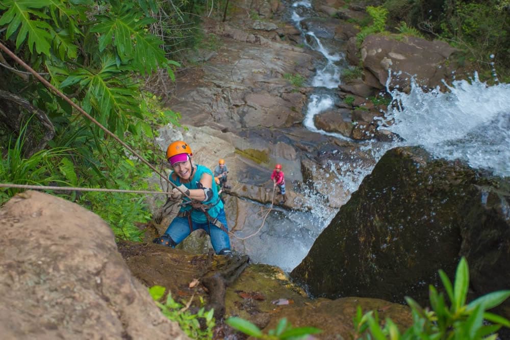 Woman repelling over edge of waterfall, in orange helmet, with water crashing around, and greenery from jungle nearby.