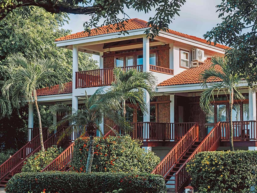 A two-story house with a red tile roof surrounded by greenery and palm trees.