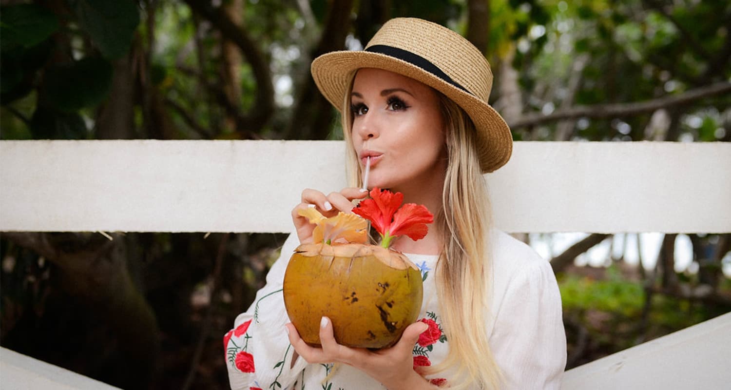 Pretty blond woman sipping in a straw hat sips on a tropical drink with an orange flower garnish.