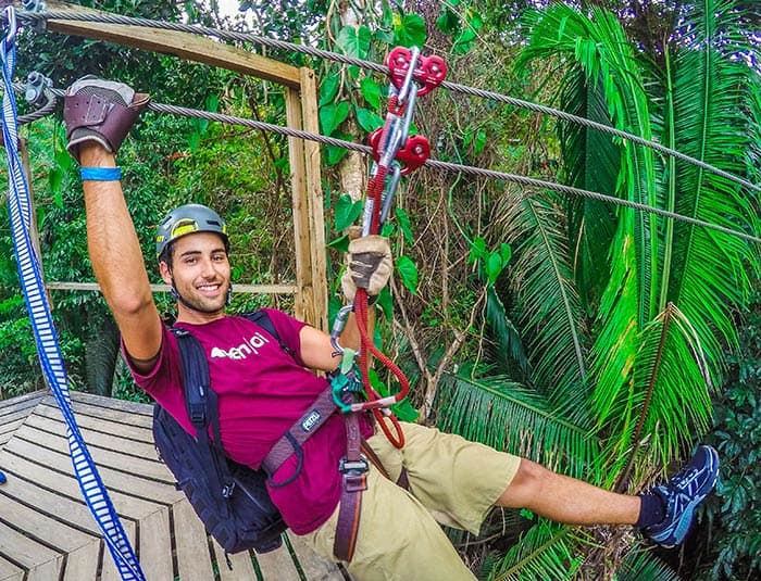 A smiling young man holds onto a zipline, getting ready to go down flying through the jungle.