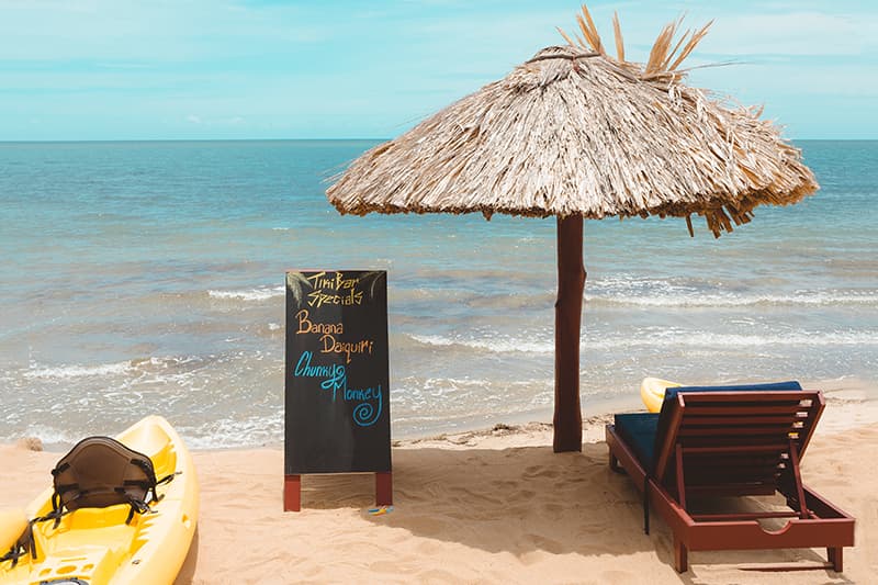A beach scene featuring a straw umbrella, lounge chair, a yellow kayak, and a chalkboard menu listing drinks. A beach scene featuring a straw umbrella, lounge chair, a yellow kayak, and a chalkboard menu listing drinks.