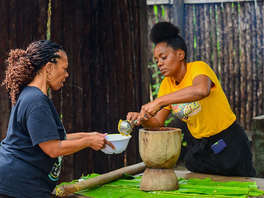 Two women are preparing food together outdoors, one mixing ingredients while the other holds a bowl.