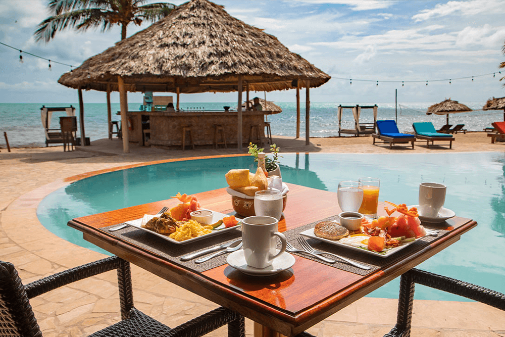 A picturesque scene of a beachfront resort in Belize. A round pool is surrounded by lounge chairs and a thatched-roof bar. A wooden table is set with a delicious breakfast spread overlooking the ocean.