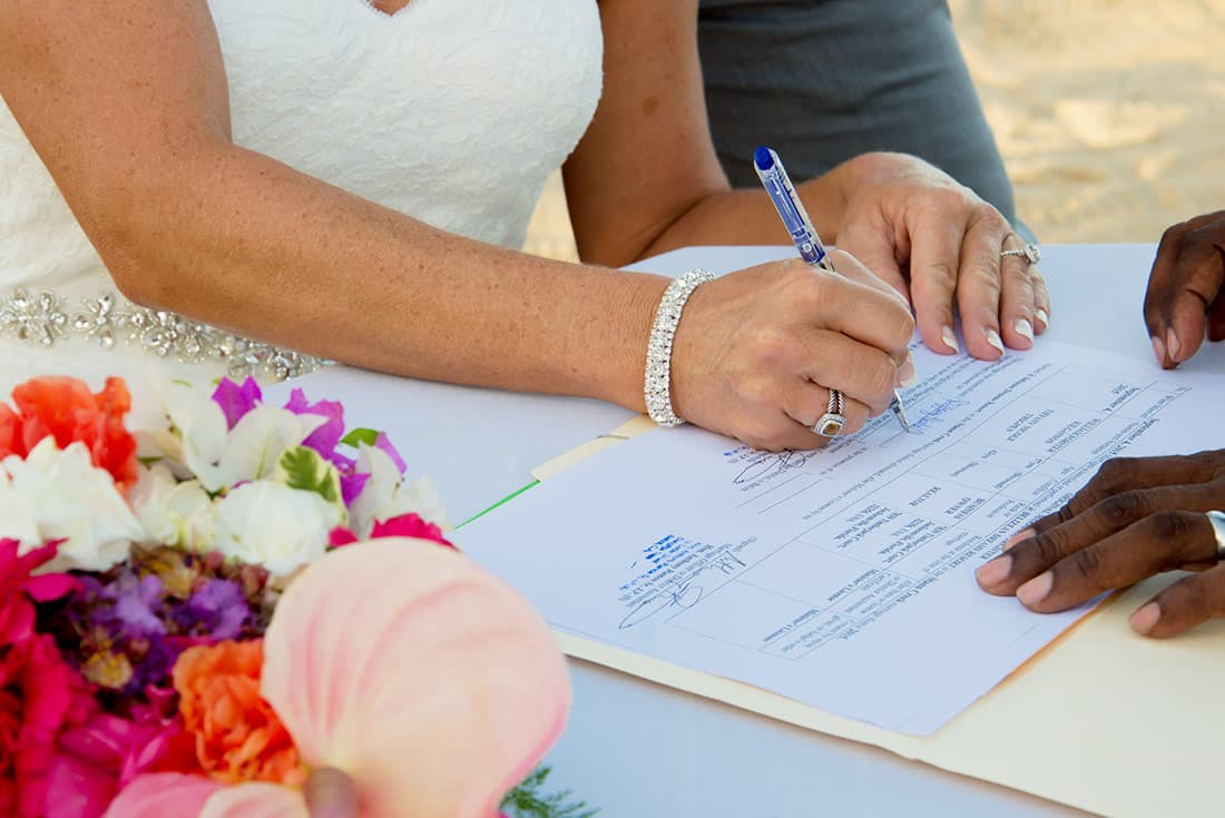 A bride in a white dress with diamond jewelry signs her marriage license next to a colorful wedding bouquet.