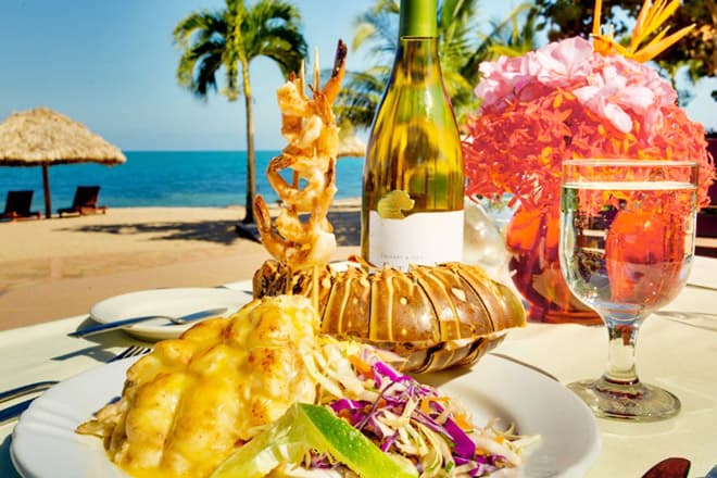 A beautifully presented seafood dish with lobster, shrimp, and colorful salad, accompanied by a bottle of white wine, set against a beach backdrop.