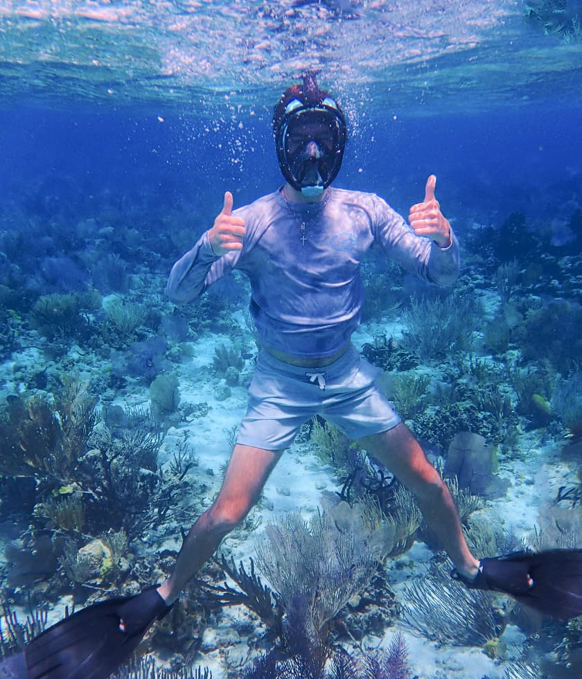A snorkeler gives two thumbs up while swimming underwater among vibrant coral and marine life.