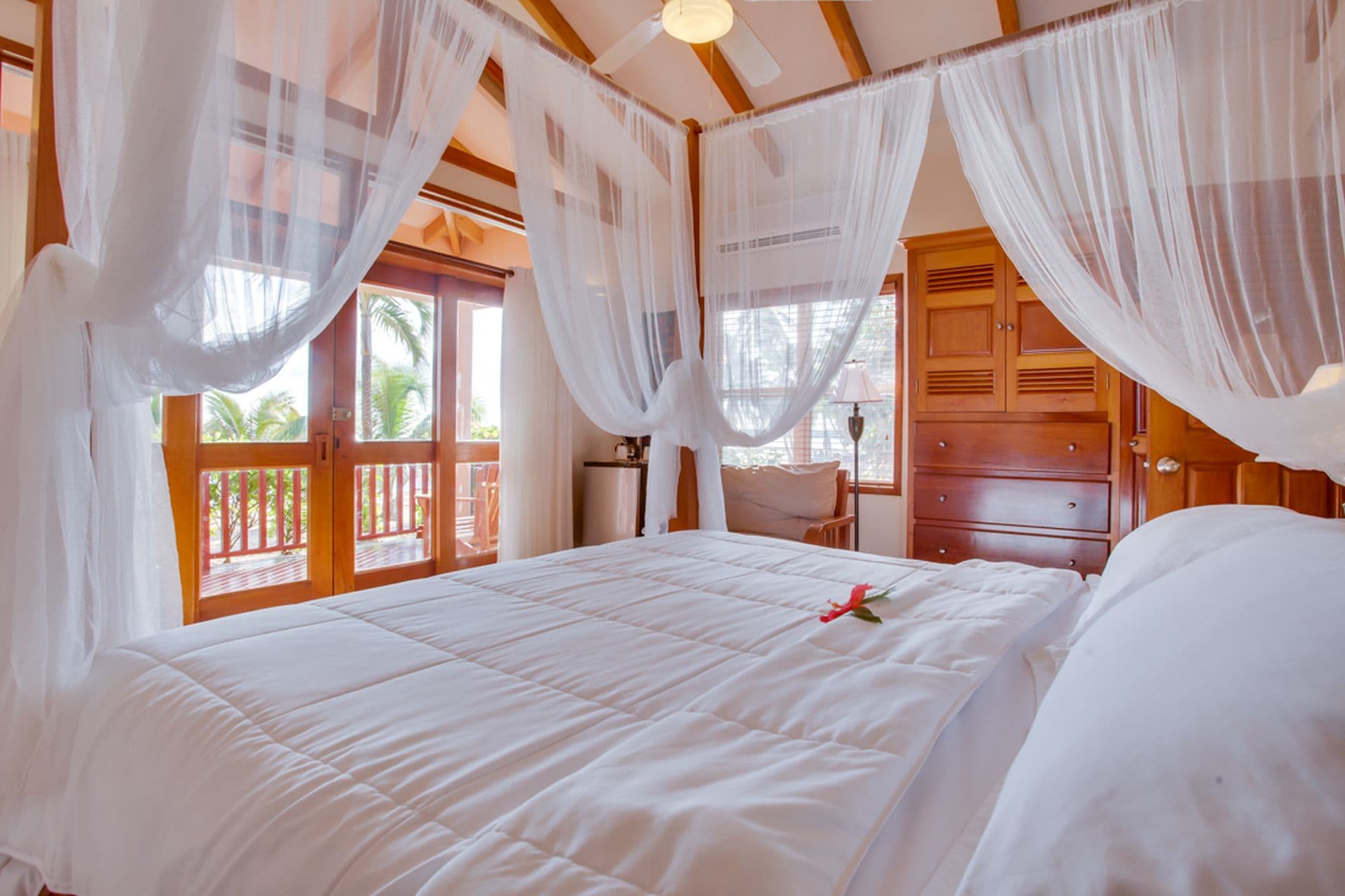 A spacious bedroom with a four-poster bed covered in white sheets. The bed has sheer white curtains hanging from the canopy. A ceiling fan hangs from the center of the room, and there is an air conditioning unit in the background. Nightstands with lamps flank the bed, and a wooden bench sits at the foot. A small sofa sits in the corner of the room, and a coffee table is placed between the sofa and the bed. Sliding glass doors lead to a balcony overlooking a view of the ocean. A colorful tapestry hangs on the wall behind the bed.