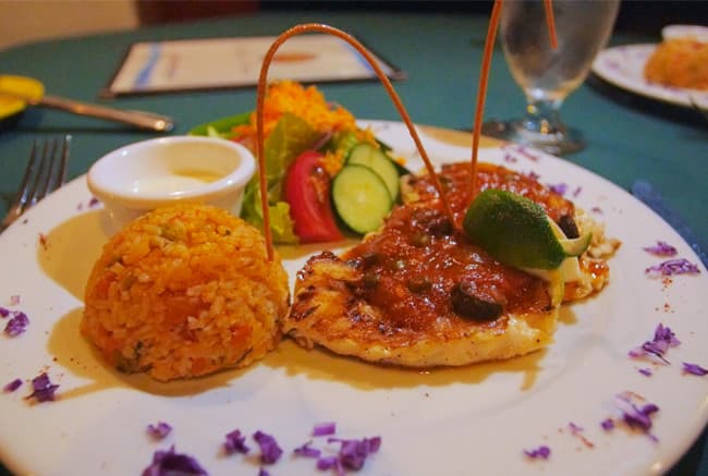 A plate of grilled chicken topped with sauce, served with rice, salad, and a side of dipping sauce.