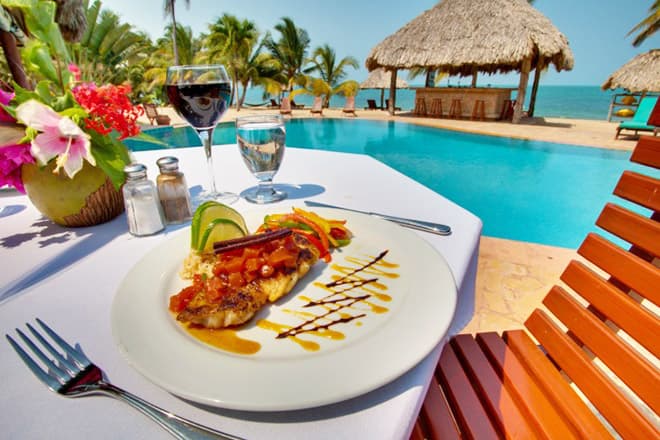 A beautifully plated fish dish with colorful garnishes and drinks sits on a table by a pool surrounded by palm trees.