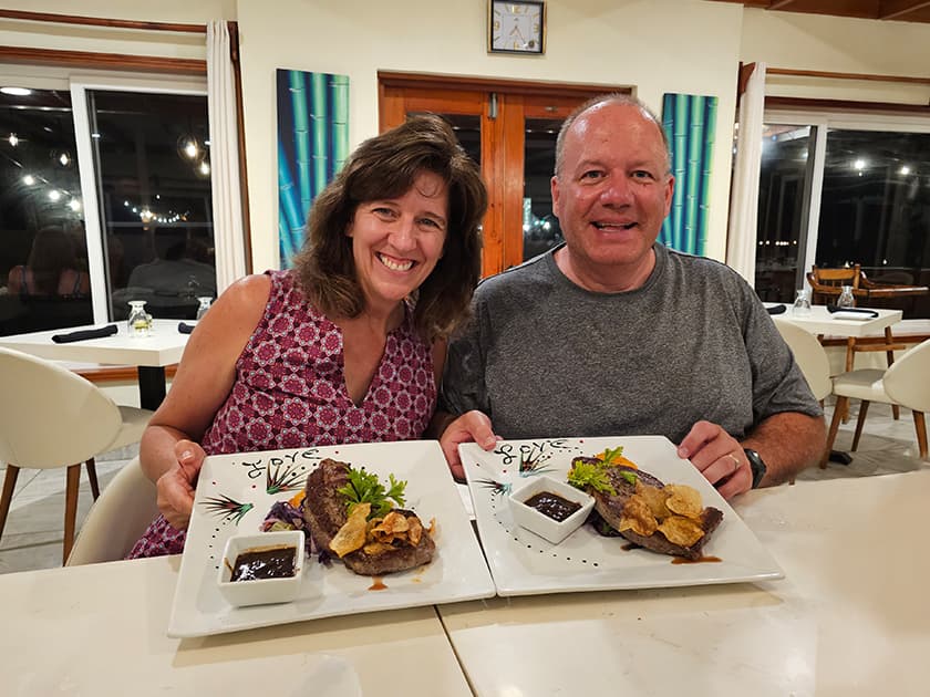 A smiling couple holds plates of steak dinner in a well-lit restaurant. A smiling couple holds plates of steak dinner in a well-lit restaurant.