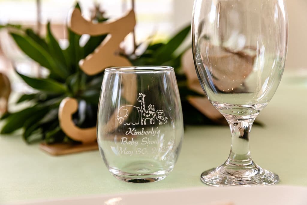 The image shows a clear glass wine glass with the text "Kimberly's Baby Shower May 30" and the silhouettes of several safari animals engraved on it. A large wooden number "3" and a water glass are visible in the background.