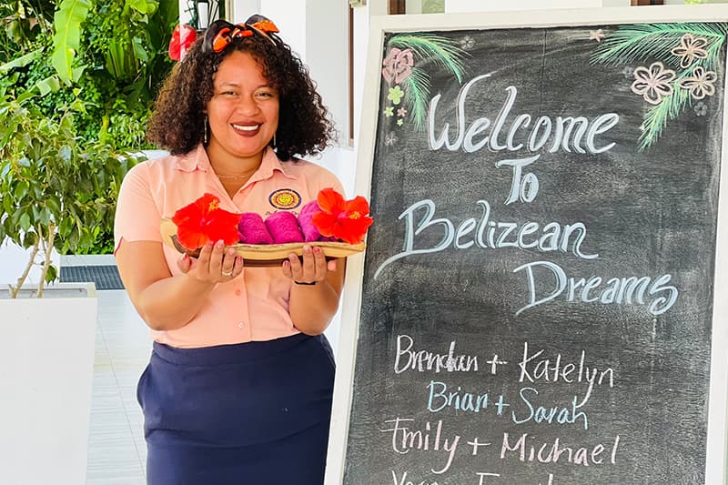 A smiling woman in a pink shirt holds a platter of fruit in front of a welcome sign for Belizean Dreams. A smiling woman in a pink shirt holds a platter of fruit in front of a welcome sign for Belizean Dreams.