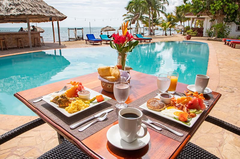 A beautifully set table with breakfast, including eggs, fruit, and coffee, by a blue pool overlooking the ocean.