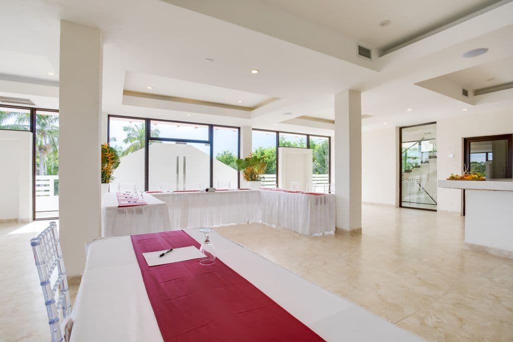 A spacious and modern meeting room with a U-shaped table arrangement. The room has a white tiled floor, white walls, and a high ceiling with exposed beams. Large windows offer views of a green outdoor space. The table is covered in white tablecloths with red runners. Clear plastic chairs are arranged around the table. There is a bar area with counter stools in the background.
