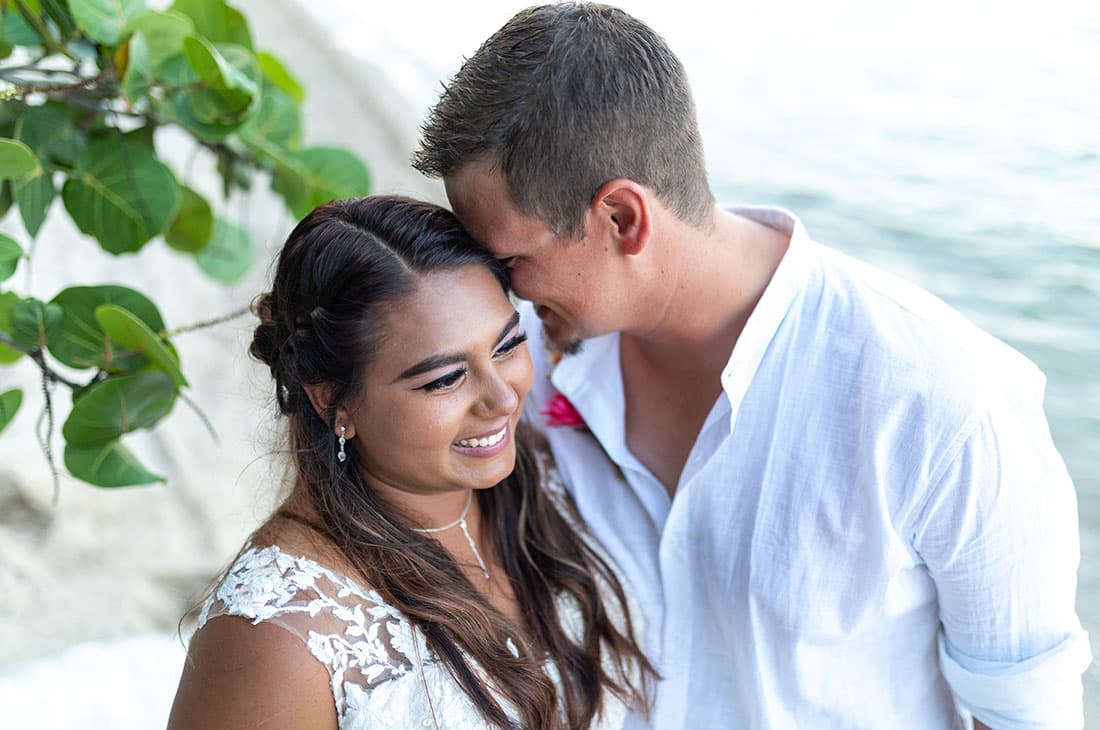 A young bride and groom in beachy wedding clothes lovingly embrace in front of a tree and the water.