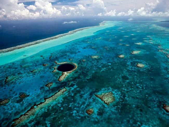 Aerial view of a vibrant blue ocean with coral reefs and a prominent dark blue underwater sinkhole.