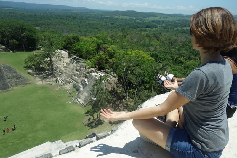 A person sits on a high ledge overlooking ancient ruins and lush green landscape.