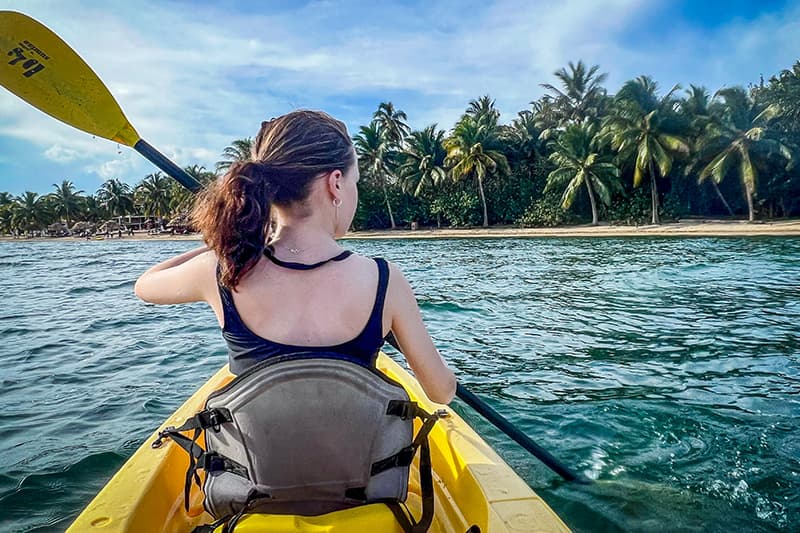 A person paddles a yellow kayak in calm waters with a tropical shoreline in the background. A person paddles a yellow kayak in calm waters with a tropical shoreline in the background.