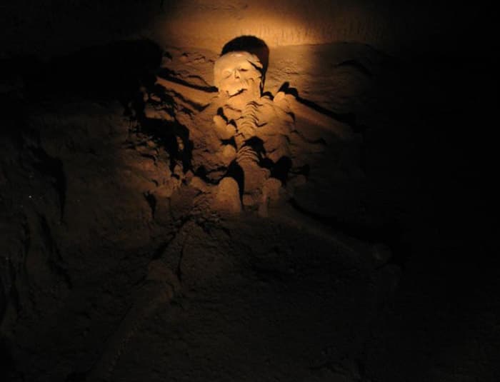 A human skeleton is partially visible in a dark cave, illuminated by a spotlight. The skeleton is lying on its side, with bones exposed.
