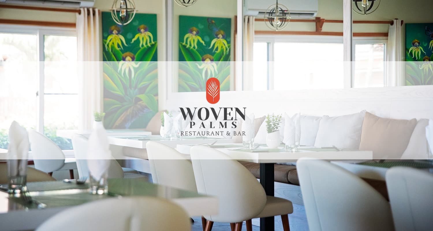 A photo of the Woven Palms Restaurant & Bar at Belizean Dreams Resort. The restaurant has a modern, upscale atmosphere with white tablecloths, comfortable chairs, and large windows overlooking the surrounding area. There are paintings on the walls and a distinctive woven palm logo above the bar.
